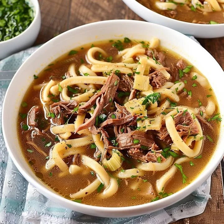 A bowl of slow-cooked beef noodle soup garnished with herbs and spices.